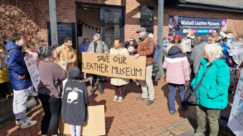 A crowd of people, mostly wearing coats, outside a brick building, with one holding a large cardboard sign with the words Save our Leather Museum on it