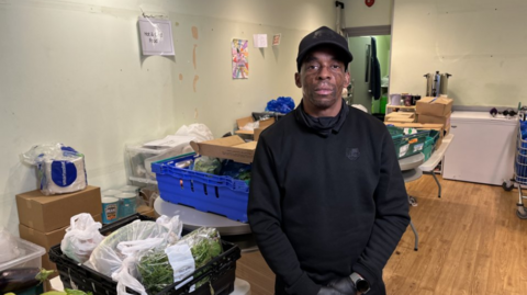 Dean Foster, a young men wearing a back cap and black clothes, standing in a room in front of boxes of fresh food