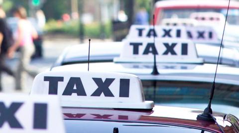 Row of taxis lined up on a city street, each with a roof sign displaying the word ‘TAXI