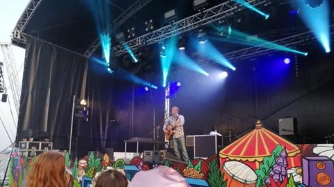 A man on stage in a patterned shirt playing an acoustic guitar and singing into a microphone. There are blue lights above him and the heads of people in the crowd are in front of the camera. The front of the stage is decorated with cartoon pictures of a red and yellow festival tent, speaks, flowers and drums.
