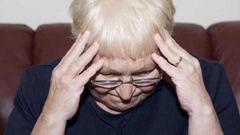 A woman wearing glasses is leaning forward holding her head in both hands.