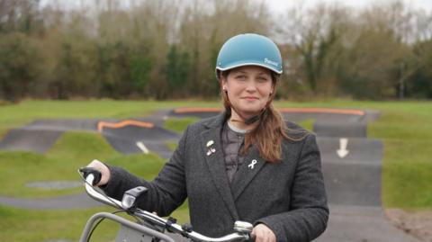 A woman with long brown hair and a grey coat wearing a blue cycling helmet and holding the handlebars of a bicycle, with a cycling track behind her