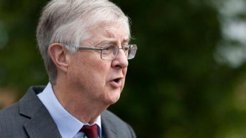 Welsh government minister Mark Drakeford wearing glasses and a red tie, speaking in front of a blurred green background