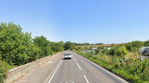 A Google StreetView screengrab showing a two lane road with a pavement on the left hand side, trees on the left and allotments on the right hand side. One white car is seen on the left hand side of the road.