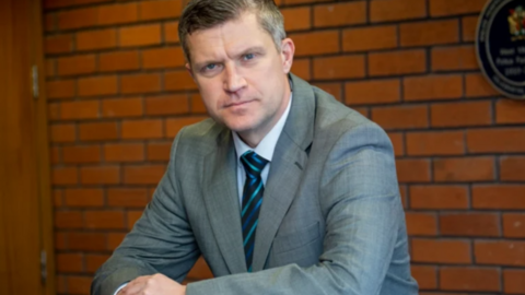 A man is wearing a grey suit, a blue tie with stripes and a shirt. He is looking at the camera and a brick wall is behind him. 
