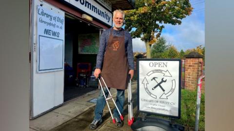 A man with blonde hair wearing a blue patterned long sleeve shirt and blue jeans, wearing a brown apron. He's holding litter pickers and standing next to a sign. 