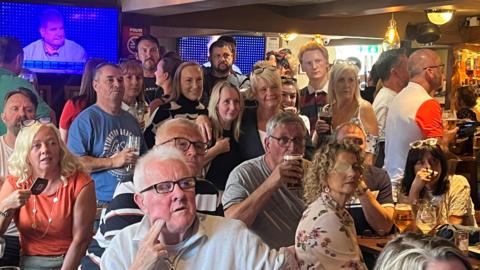 Inside the Colliers pub in Tyldesley packed with friends and family drinking and watching intently the late Tim McCarthy on the Chase. Tim McCarthy is shown on a TV screen in the background taking part in the show. 