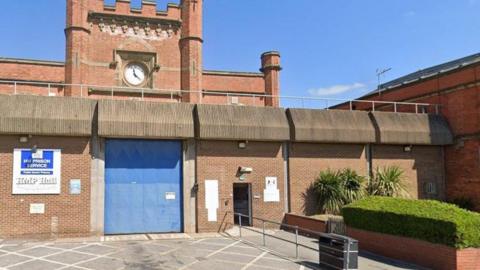 The outside of HMP Hull, as denoted by a sign on a wall. A large blue entry door, which is closed, is visible, with high walls on either side. A clock tower protrudes from behind.
