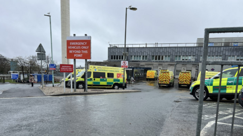 Ambulances parked in a car park outside of a hospital. The hospital building is large and grey. A large red sign is displayed in front of the car park which reads 'emergency vehicles only beyond this point'. 