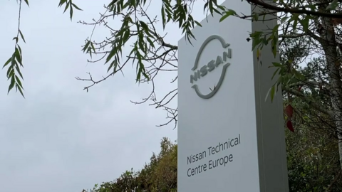 A white sign with the silver Nissan logo, and the words "Nissan Technical Centre Europe". It is next to a hedge and a tree.