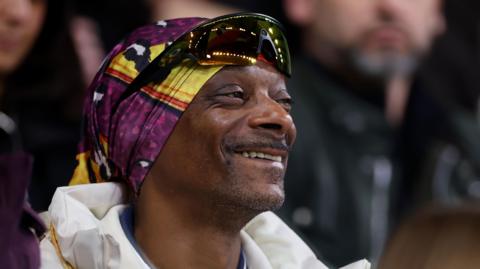 Snoop Dogg at a Swansea football game wearing a white Swansea FC jacket and t-shirt. He also wears a colorful bandana and yellow sunglasses perched on top of his head. He is smiling. 