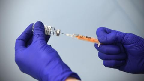 A vaccine is extracted from a vial using a syringe, each object is held by a hand in blue gloves.