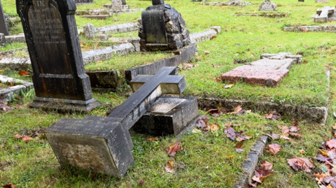 A grave headstone that has been laid flat, the headstone is in the shape of a cross and has been placed against the base of the grave. It is pictured on grass which is partially covered with autumn leaves.