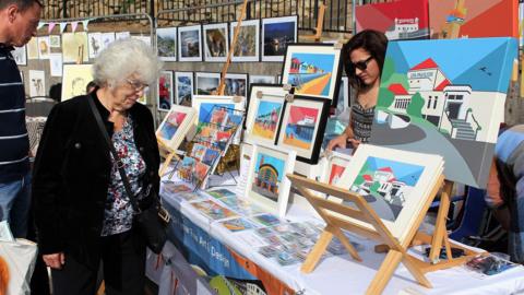 A woman browses a stall an an open-air art market. A woman with black hair and sunglasses stands behind the stall.