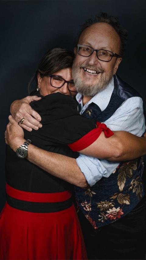 Lili Myers and Dave Myers stand closely together in a warm embrace, both smiling. Lili wears glasses, a black top with red accents, and a red skirt. Dave also wears glasses along with a light blue shirt and a dark floral-patterned vest. The background is dark, drawing focus to their affectionate pose and expressions.