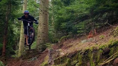 A mountain biker is flying mid-air while he completes a trick. The photograph was taken in Coombs Wood, Armathwaite.