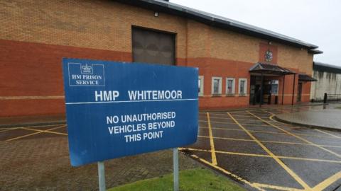 A blue sign saying HMP Whitemoor in white letters outside a yellow and orange brick building.