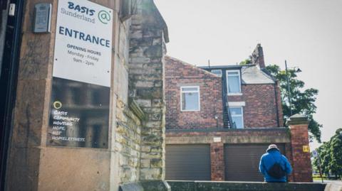 The entrance to a stone building, which has a sign that says 'Basis' on the front of it. To the right of the building is a man in a blue coat.