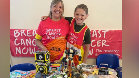 Woman with shoulder-length blonde hair smiling with a red Breast Cancer Now t-shirt. Next to her is a boy with short brown / blonde hair with a black t-shirt and a Breast Cancer Now tabard. There is a red Breast Cancer Now banner behind them on a beige wall. In front of them there is a table with toys on it and there are also two blue chairs. 