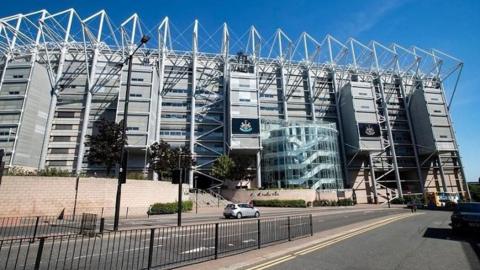 St James' Park is a large metal structure with Newcastle United's crest on two of the panels. Barrack Road, immediately outside it, is a long road with four lanes. A bus and a car are travelling on it. 