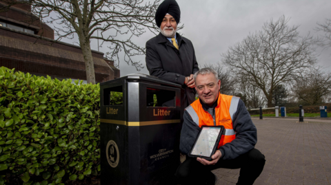 Councillor Bhupinder Gakhal, cabinet member for resident services at City of Wolverhampton Council, with Andy Moore, public realm team leader. They are by a litter bin in the city with Mr Moore holding some of the technology linked to the sensors. Mr Gakhal is leaning on the bin with Mr Moore crouched beside it.