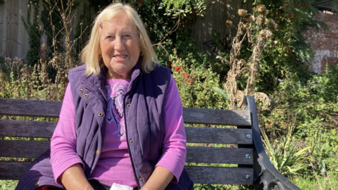A woman with blonde shoulder length hair, wearing a purple vest over a pink top sits on a wooden bench in a sunlit garden, surrounded by green plants and trees, with some dried vegetation in the background.