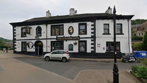 The exterior of a large building set on the side of street. The building is painted in white and black. On the front is a sign in gold letters which reads 'The Golden Lion'.