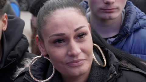 Victoria Phillips has scraped back hair and big hooped earrings. She is looking away from the camera and giving an interview. There are people behind her. 