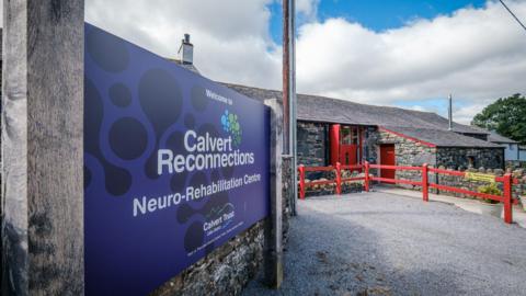 A building sign on the left reads: "Calvert Reconnections Neuro-Rehabilitation Centre". The single-storey building on the right is made from stone and has painted red doors and fences.