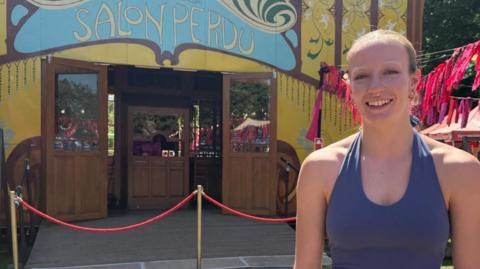 Chelsea stands in front of the Norfolk and Norwich Festival's yellow circus tent on a sunny day. She is smiling, wearing a purple halterneck leotard. Her hair is blonde and pulled back from her face.