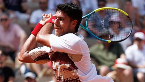 Adolfo Daniel Vallejo, wearing a white patterned t-shirt, plays a backhand with his yellow, blue and black racquet held in two hands over his right shoulder