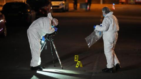 Two crime scene investigators, dressed in hazmat suits, is conducting an investigation on a road. One of them is taking photographs while the other one is holding a bag to store crime scene evidence. 