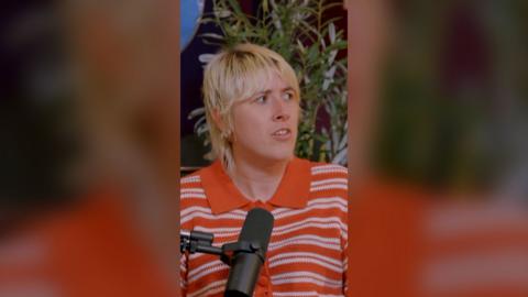 Maisie Adam looks to her left. She's wearing an orange striped polo shirt and has a serious expression on her face.