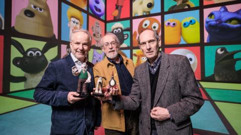 Aardman animation's co-founder and creators are standing in front of screen showing a collection of the studios most famous characters, including Wallace & Gromit, Shaun the Sheep and Morph. Peter Lord is standing in the middle wearing a brown Jacket holding a Gromit figurine. Nick Park is standing to the left of him wearing a navy blazer and holding a Wallace figurine and David Sproxton is standing to the right of Peter holding a penguin figurine. 