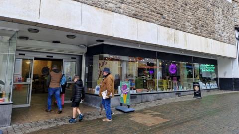 The front of a shop with various illuminated signs in the window. Two men wearing brown jackets and blue jeans, and a boy in shorts and a black coat are walking into the open double doors. There is a sign outside advertising Gelato ice cream.