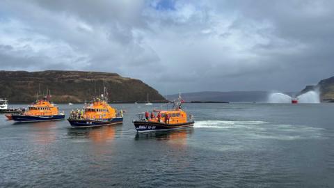 Three orange and blue coloured lifeboats can be seen travelling through a narrow bay. Behind them in the distance is a larger coastguard ship spraying its water cannons. 
