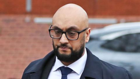 A man with a bald head and wearing glasses, a blue overcoat, white shirt and blue tie looks off camera. Behind him is a brick wall.