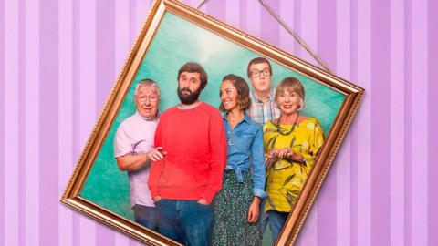 A crooked picture frame hangs on a pink wall. In the image a family is shown, wearing bright colours.