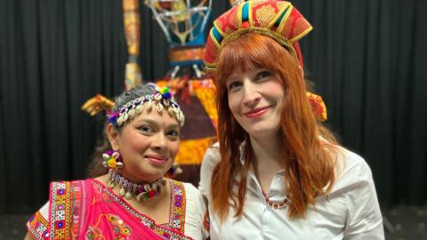Two women stand arm in arm smiling. The one on the left wears and headdress and necklace made of shells and colourful thread and the one on the right wears a headdress made of red and gold satin. 