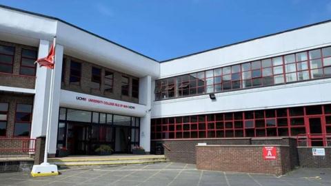 The exterior of University College Isle of Man, known as UCM. The building is white, with brown bricks and red rimmed windows, and there is a red Manx flag on a flagpole in front of it.