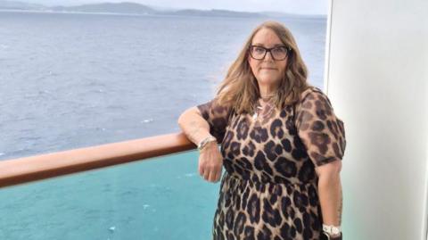 Emma is standing next next to the ocean with her arm rest on a glass ledge. She is wearing glasses and a leopard print dress. 