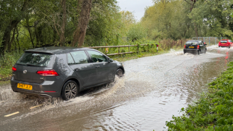 Three cars driving through floodwater on a country road. The water looks to be several inches deep. Either side of the road is greenery, bushes and trees.