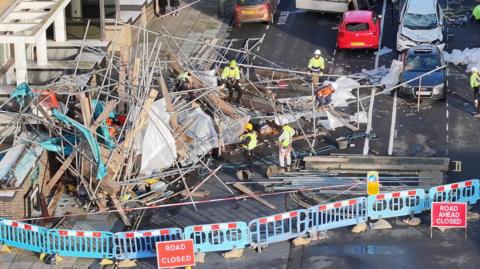 An aerial view of a huge pile of scaffolding poles and wooden planks blocking First Avenue in Hove, which is cordoned off with blue plastic barriers and road closed signs. Workmen in yellow hard hats and high vis jackets are starting to clear the mess. Damaged cars can be seen in the background.