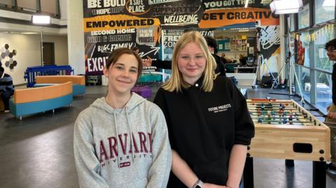 Two girls smiling at the camera as they pose for a picture in a youth club. Maisie on the left has brown hair pulled back in a pony tail and is wearing a grey hoodie with Harvard University branding in large red type. Iga on the right wears a black hoodie bunched up on her arms. She has shoulder-length blond hair and has rimless glasses.