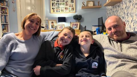 A family of four sit on a sofa in a cosy, busy living room. All are smiling at the camera. On the left is a woman wearing a pale blue jumper. A young girl and a young boy sit next to her with a man on the right wearing a beige jumper.