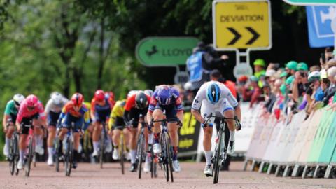 A tight knit group of about a dozen racing cyclists. To the left, spectators line the road behind a barrier and in the background, sightly out of focus, trees can be seen with signs saying "deviation" and "Lloyds" (the sponsors).