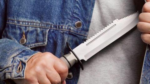 A stock image of a person holding a large silver and grey zombie knife. The picture is a close up of their hands and torso, and they are wearing a grey top and blue denim jacket. 