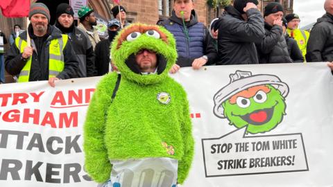 Protestors stand outside Coventry Council House. One protestor wears a green monster costume next to a banner which reads "Stop the Tom White Strike Breakers"