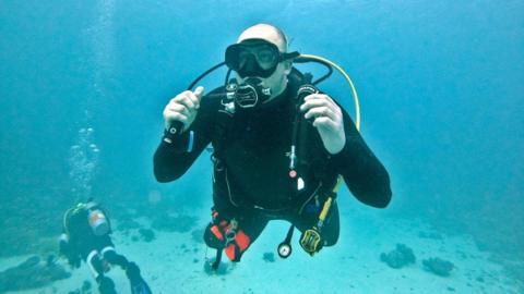A man is wearing a full scuba diving suit and equipment and is underwater in clear blue water.