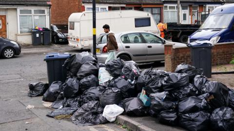 A large pile of black bin bags on the side of a residential street in Birmingham. A pedestrian is walking past the bags, and there are various vehicles parked on both sides of the road.